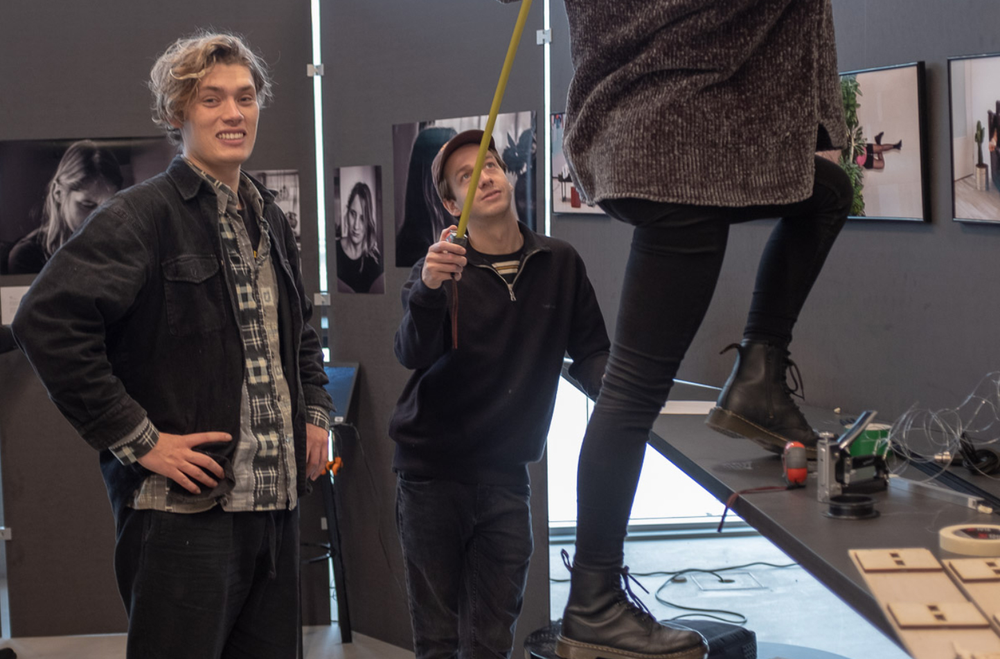 Studenten Fotojournalistiek staan in de tentoonstellingsruimte met foto's aan de muren en zijn bezig met de opbouw. Eén student kijkt lachend in de camera, een tweede houdt een meetlint omhoog en de derde staat op een krukje en is daarom alleen voor de onderste helft te zien.